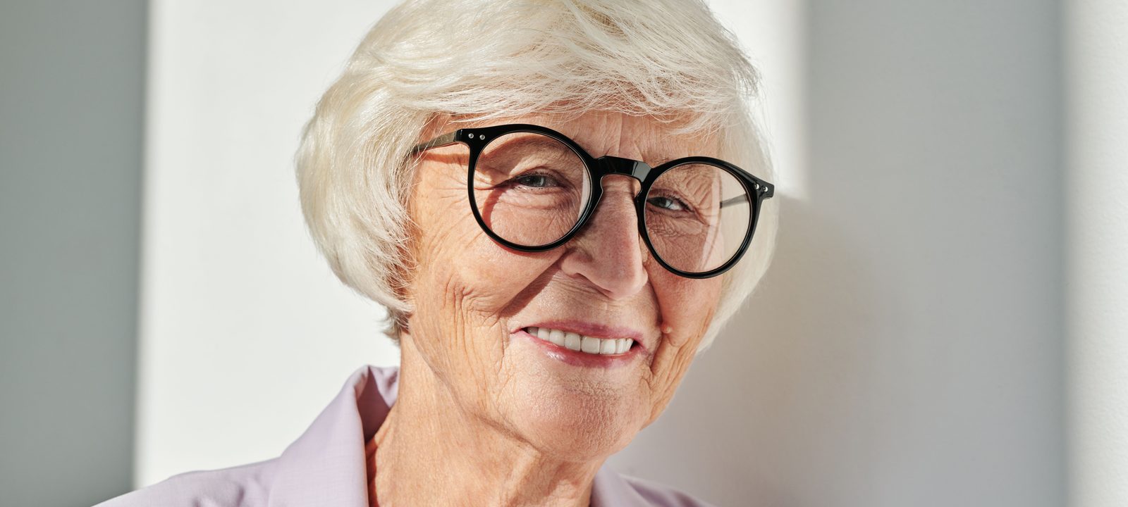Close-up portrait of a smiling senior woman with glasses enjoying the sunlight indoors.
